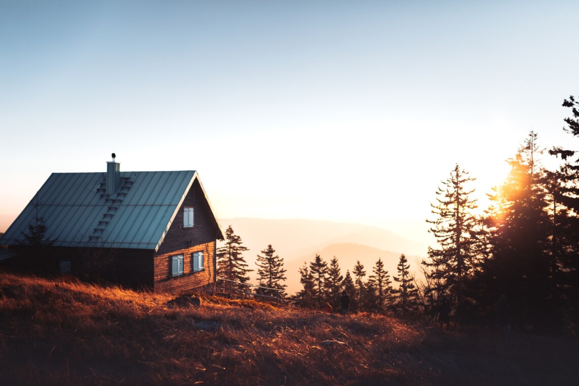 A cabin sits amongst trees on a mountainside, with the sun setting in the background