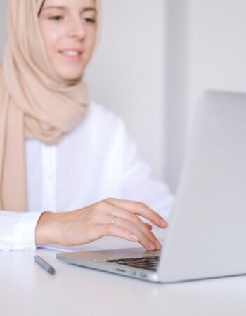 A woman wearing a hijab sits smiling at her laptop