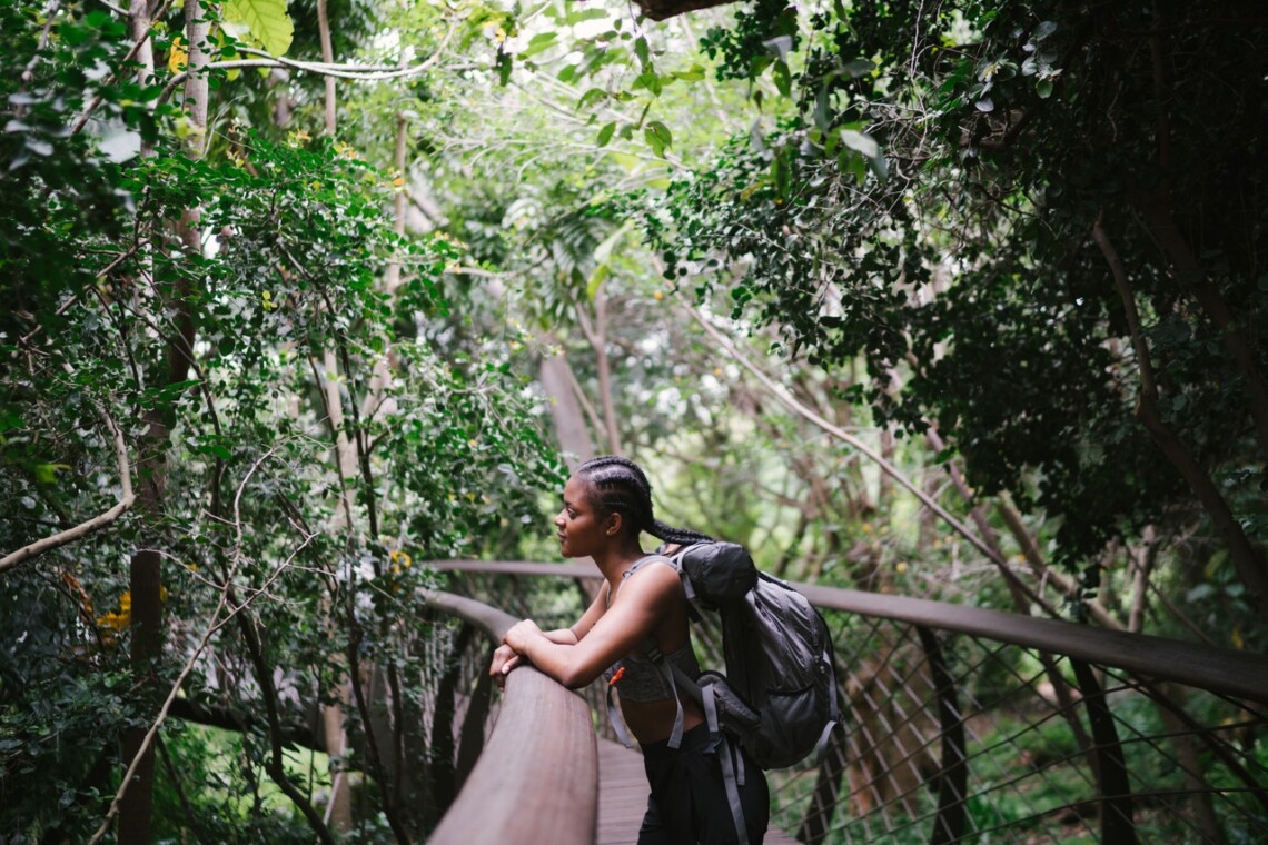 A Black woman stands on a bridge that curves through a forest. She is wearing a backpack and looking thoughtful