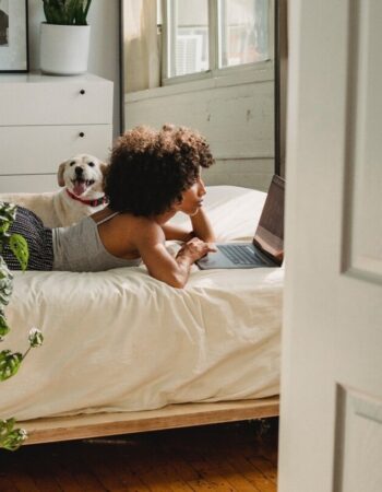 A Black woman lays on a bed with a small dog next to her. She is on her laptop, and her room is full of houseplants.