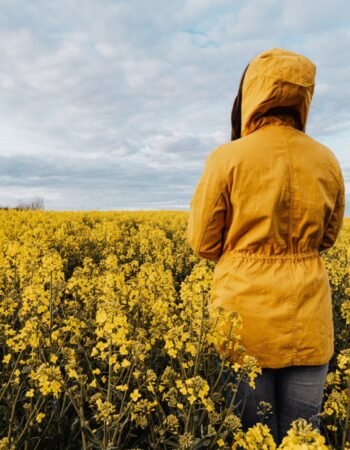 A woman in a yellow jacket stands in a field of yellow flowers, facing away from the camera, looking thoughtful