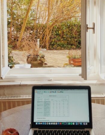 A laptop sits on a wooden table in front of an open window.