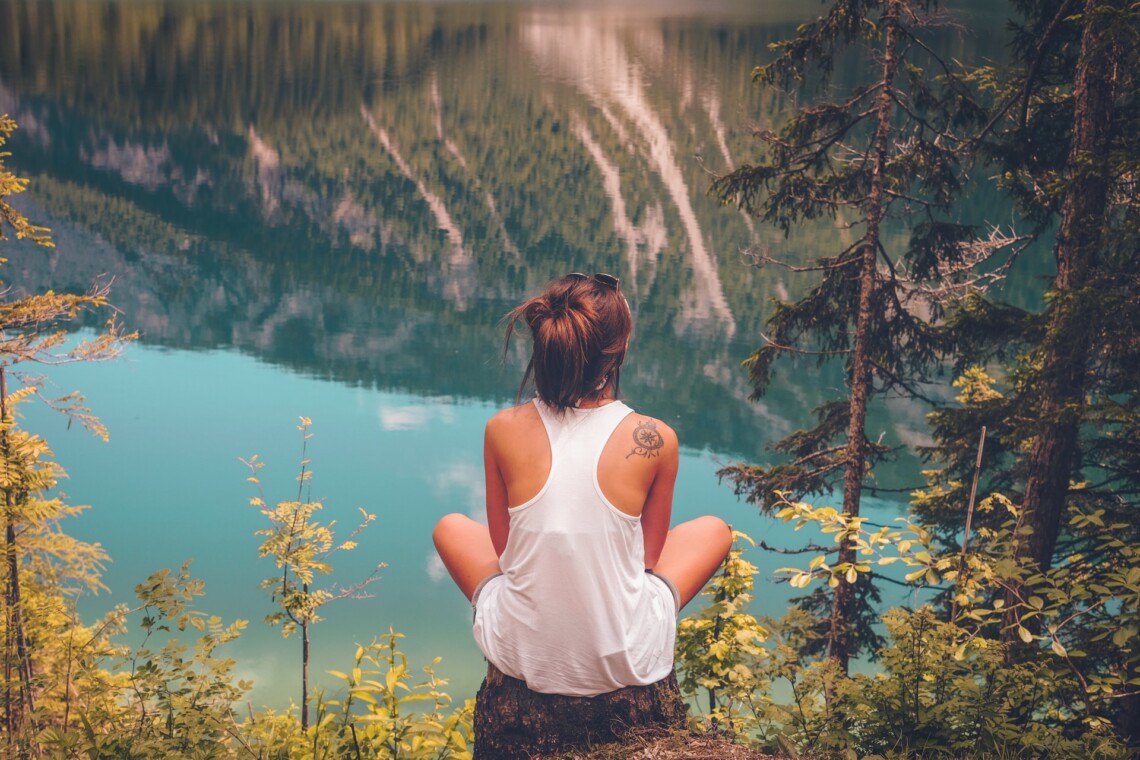 A woman sits with her back to the camera on a tree stump looking out over a lake and mountains. She looks thoughtful.