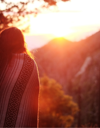 A woman stands with her back to the camera, wrapped in a blanket. There is a bright red sunset over mountains in the background