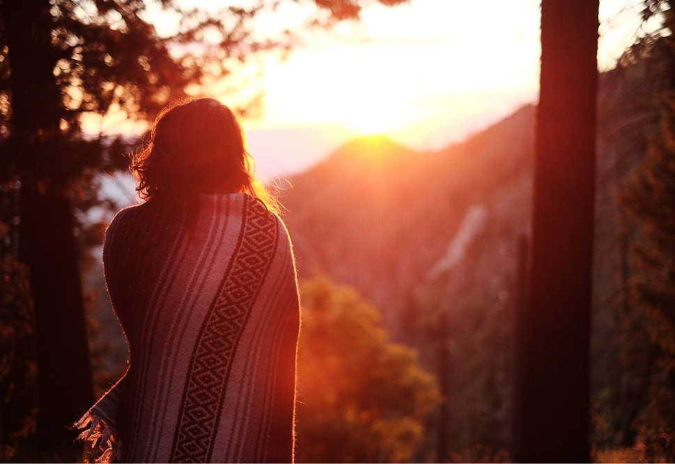 A woman stands with her back to the camera, wrapped in a blanket. There is a bright red sunset over mountains in the background