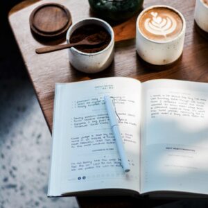 In the foreground, a notebook covered in writing sits open with a pen on the page. In the background there is a beautiful coffee