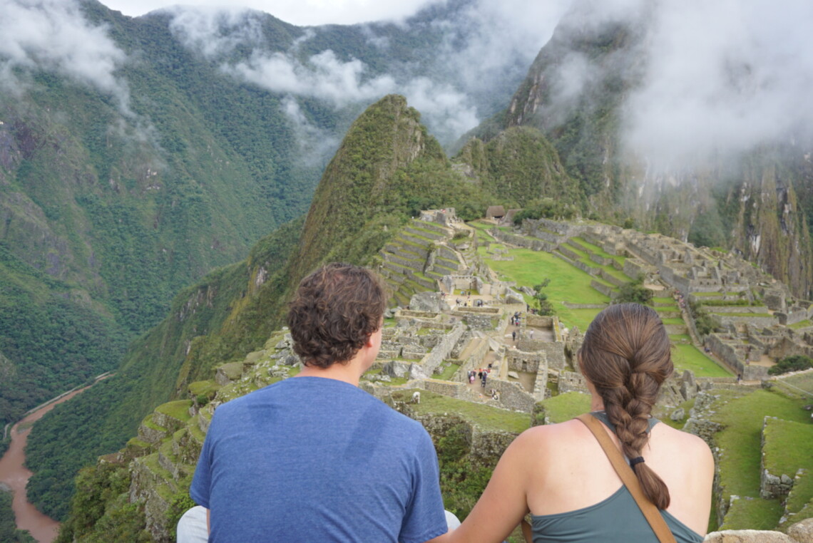 A woman and man (the ones who run this website) have their backs to the camera and are looking out at Machu Picchu. It's foggy and the clouds sit on top of the green mountains above the Incan ruins