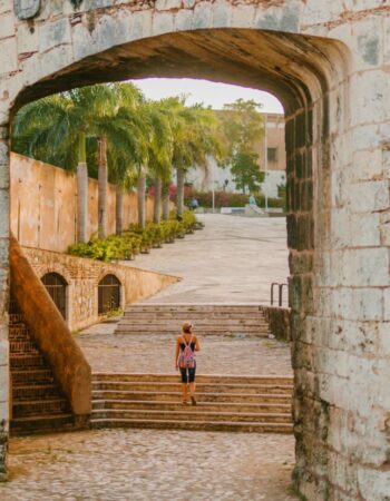 A woman walks underneath of an old, aged stone archway up steps. There are palm trees in the background and the image inspires adventure and travel.