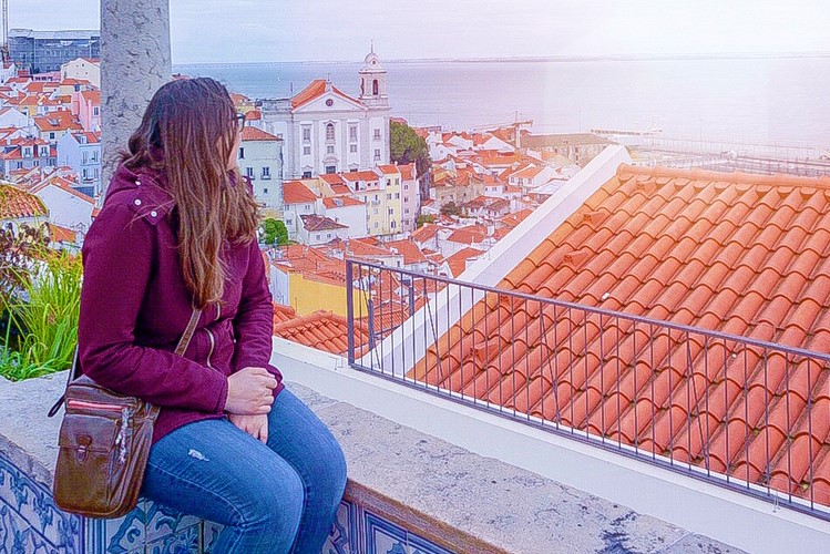 A woman sits with her face away from the camera, looking out at a viewpoint over red tiled rooftops and water