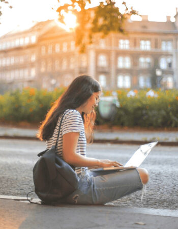 Woman sitting on her laptop outside writing at sunset wearing a backpack