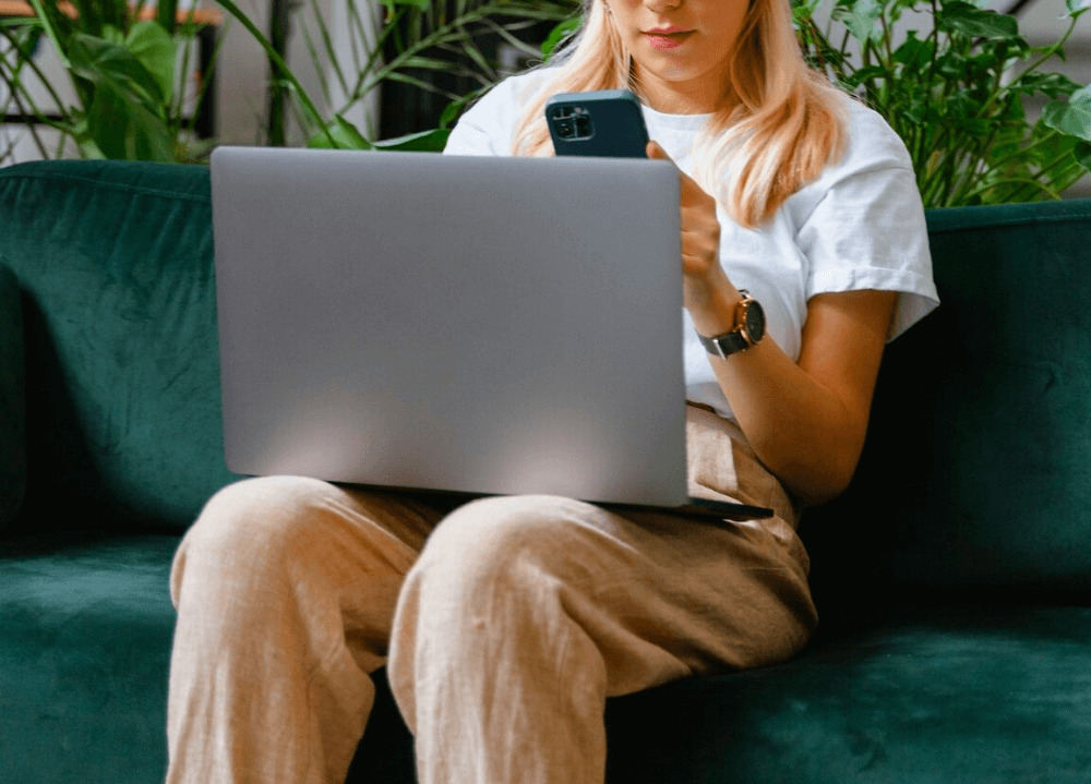 A woman is sitting on a couch with a laptop on her lap while looking thoughtfully at her phone