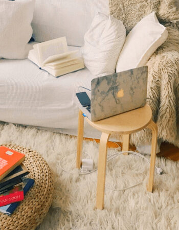 A Macbook sits on a small table next to a cozy couch with books on the coffee table