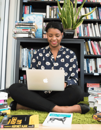 A Black woman sits on the floor in front of bookcases on her laptop, smiling, surrounded by books on the floor