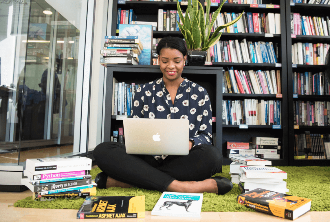 A Black woman sits on the floor in front of bookcases on her laptop, smiling, surrounded by books on the floor