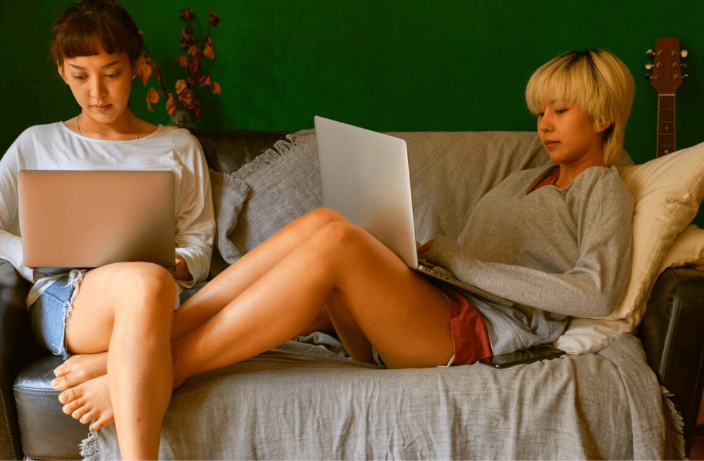 Two young women sitting on a couch together on their laptops