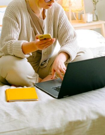 A woman sits on a bed with her laptop, phone and notebook looking very busy