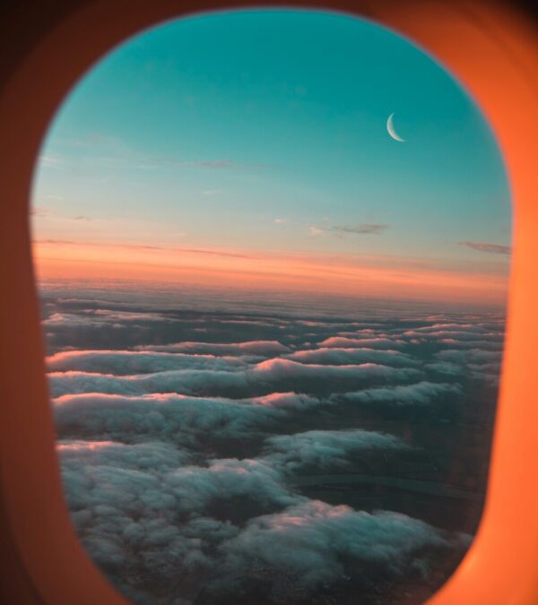 The view from an airplane window at sunset. The clouds and plane window are lit up pink from the sunset, and a crescent moon hangs above the clouds
