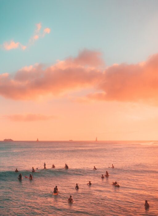 Surfers sit on the surf boards waiting for a wave. The water and sky are a beautiful pink and blue, very dreamy
