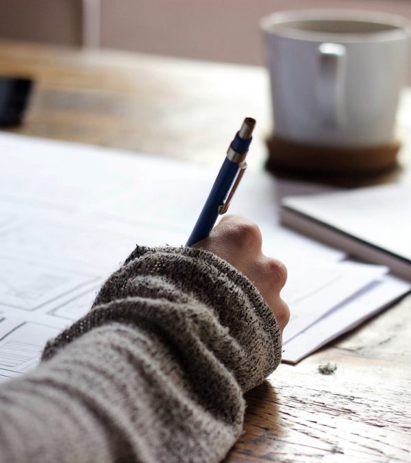 A woman's hand is writing on paper. There is a cup of coffee and the background is blurred
